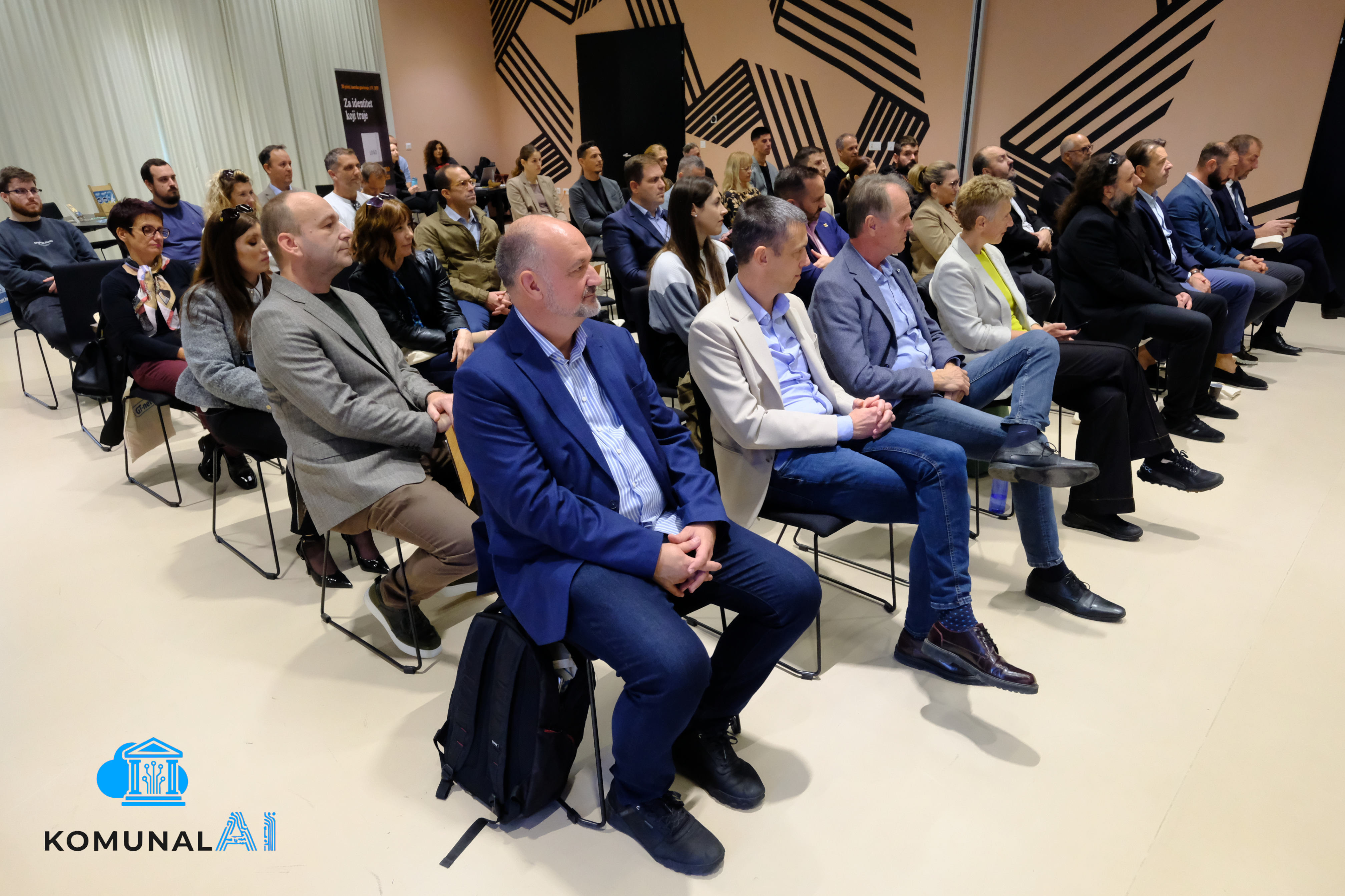 Raznovrsna publika u modernoj dvorani: muškarci i žene u poslovnim odijelima sjede na stolicama, slušaju predavanje.