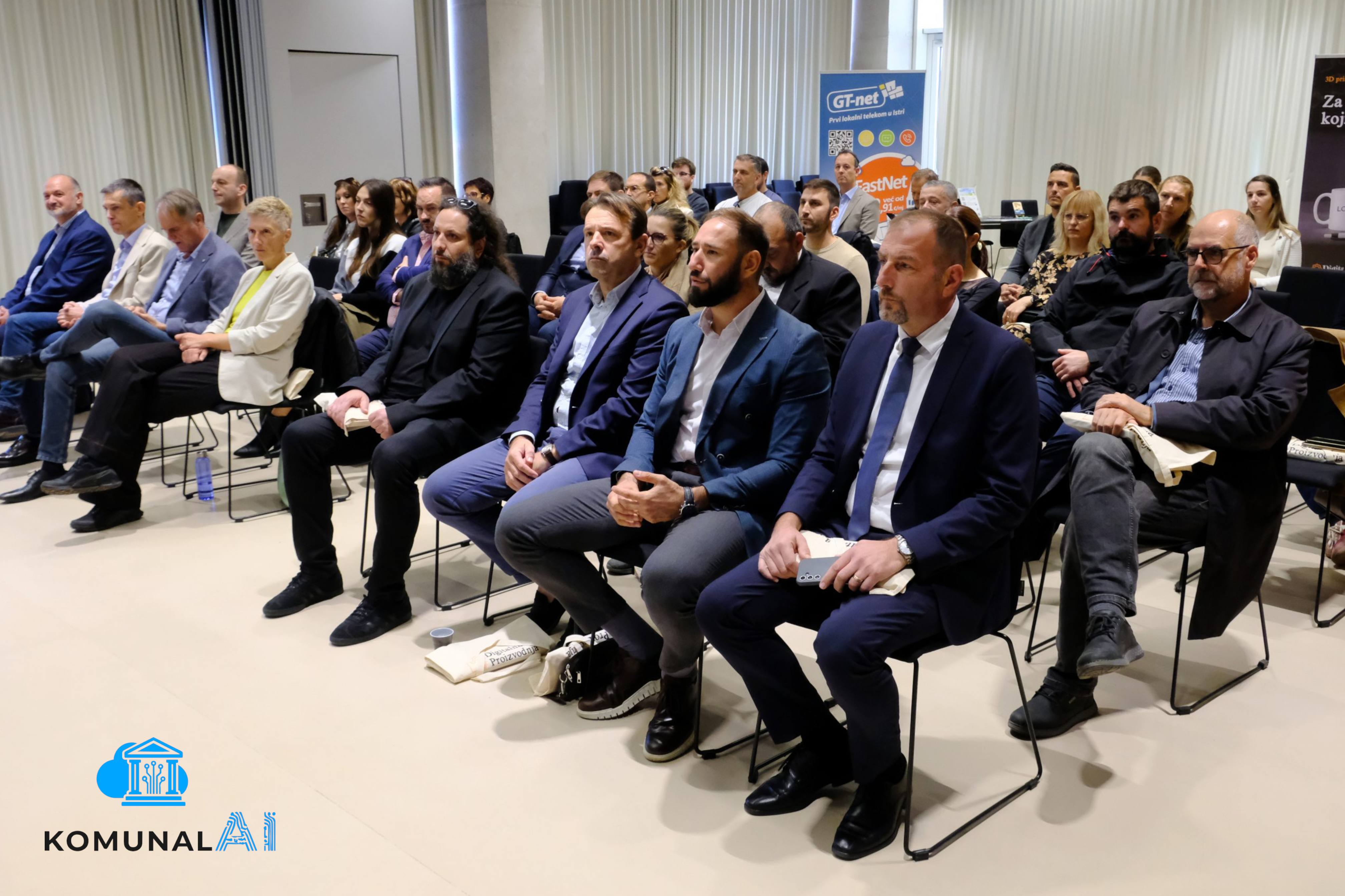 Fotografija prikazuje publiku u konferencijskoj dvorani tijekom događaja o digitalizaciji javne uprave. Među sudionicima su predstavnici GT-neta, lokalnog telekom operatera, te stručnjaci i djelatnici iz sektora javnih i komunalnih usluga. Svi pažljivo prate prezentaciju, dok se u pozadini vide promotivni materijali GT-neta i vizualni elementi povezani s telekomunikacijama i tehnologijom.